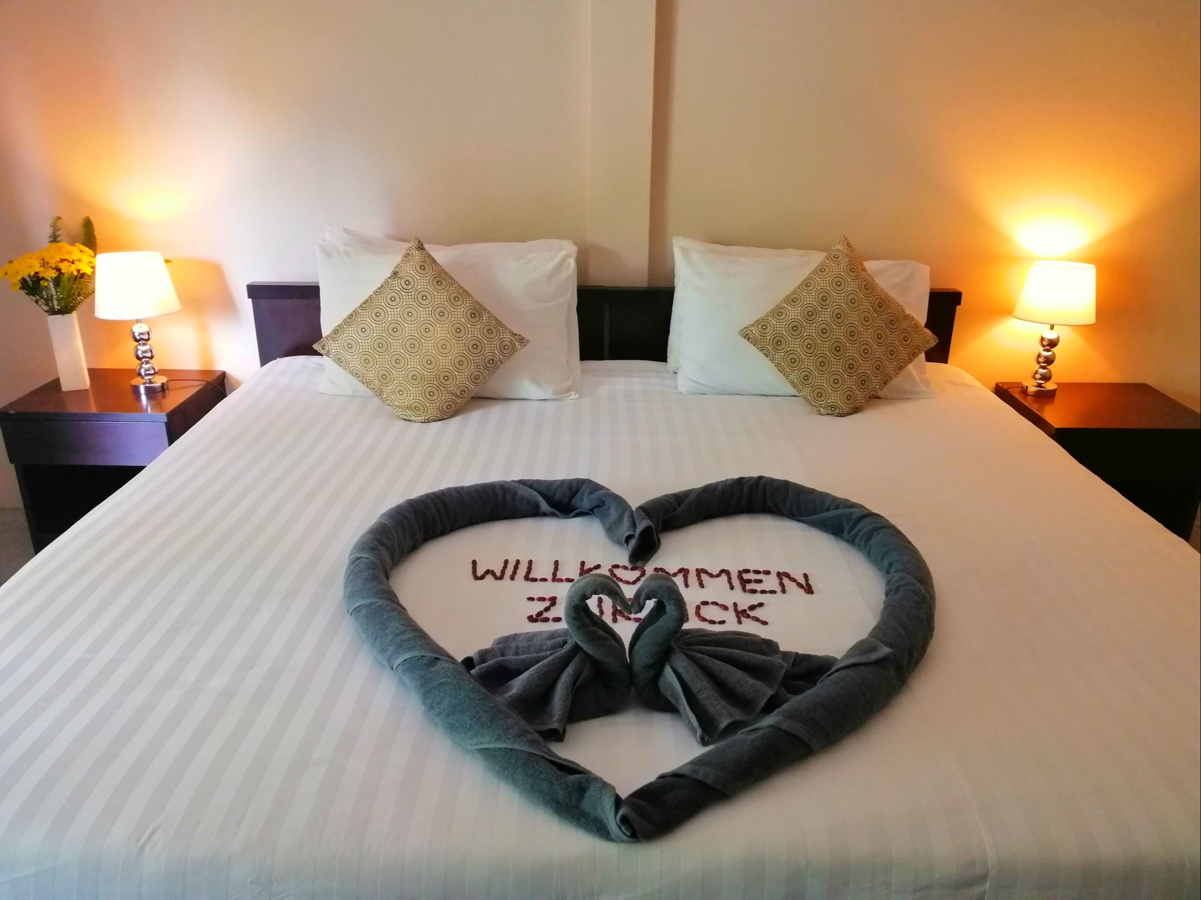 A hotel room with a neatly made bed, two bedside lamps, and a heart-shaped pillow with "WILL KOMMEN ZUCK" written on it.