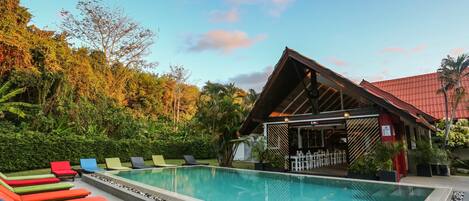 A swimming pool with lounge chairs, surrounded by greenery and a building with a red roof.