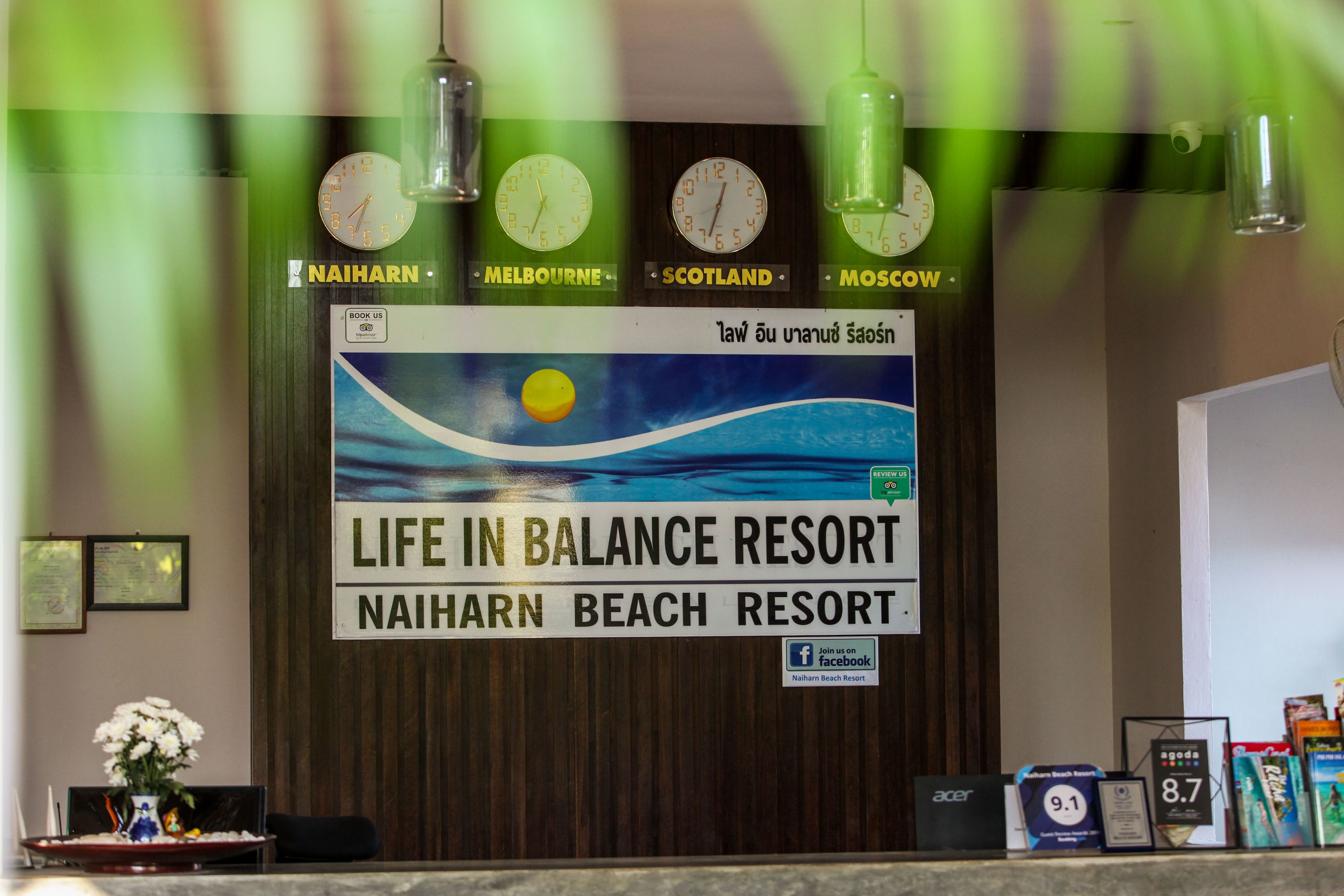 Een receptiebalie bij Na'iharn Beach Resort met klokken die verschillende steden aangeven, een bord met de naam van het resort, en een Facebook logo.