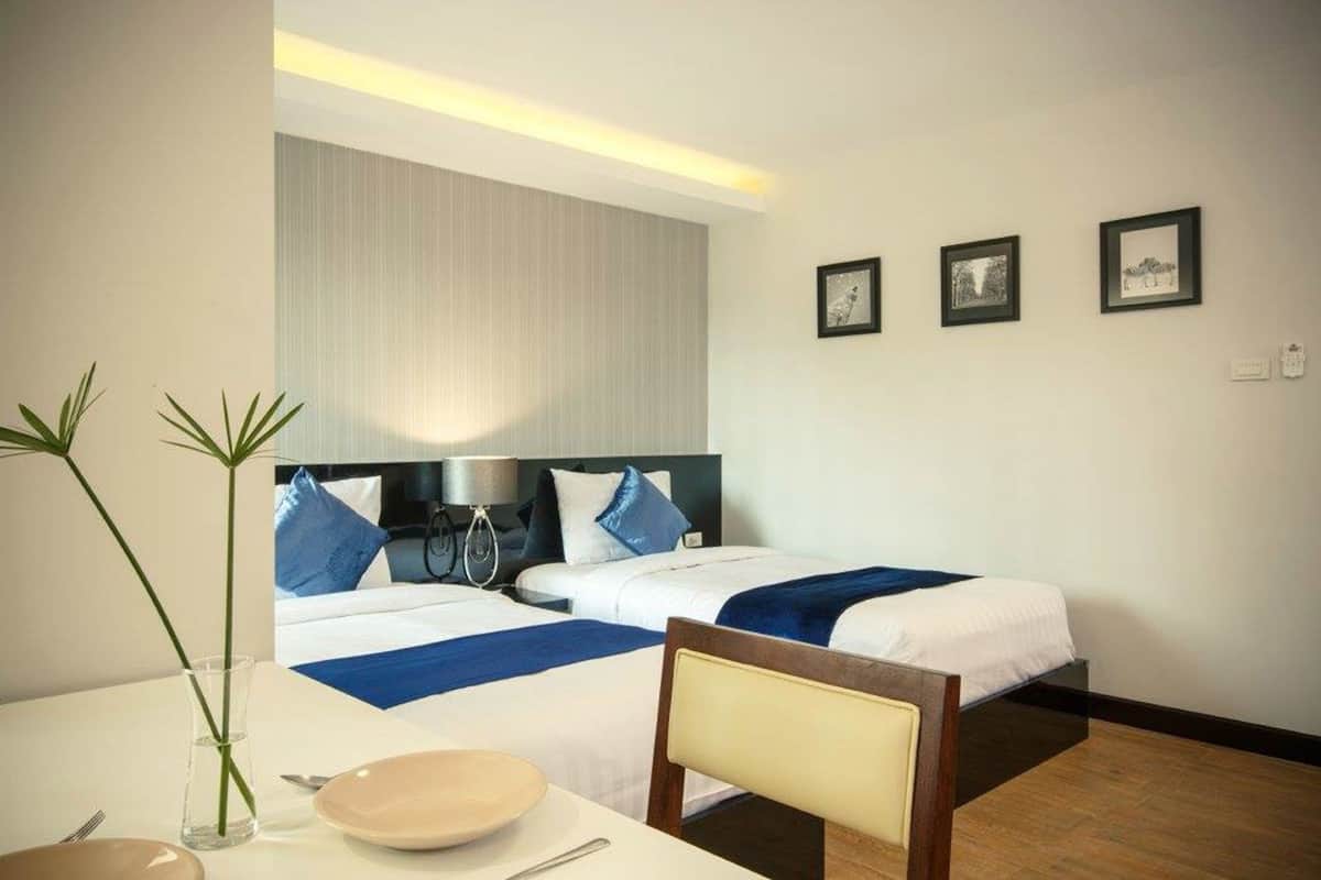 A hotel room with two beds, a dining table with a plate and a vase, and three framed pictures on the wall.