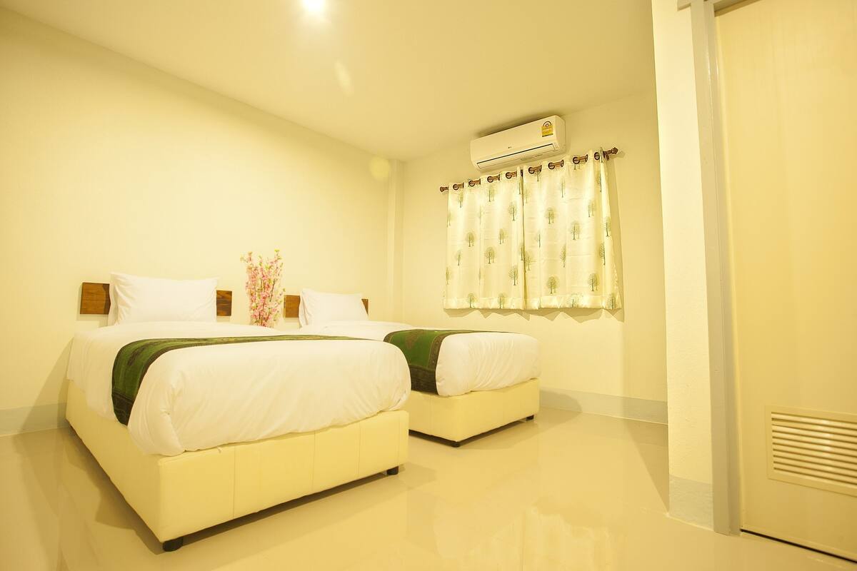 A hotel room with two beds, a ceiling-mounted air conditioner, and a window with a curtain.
