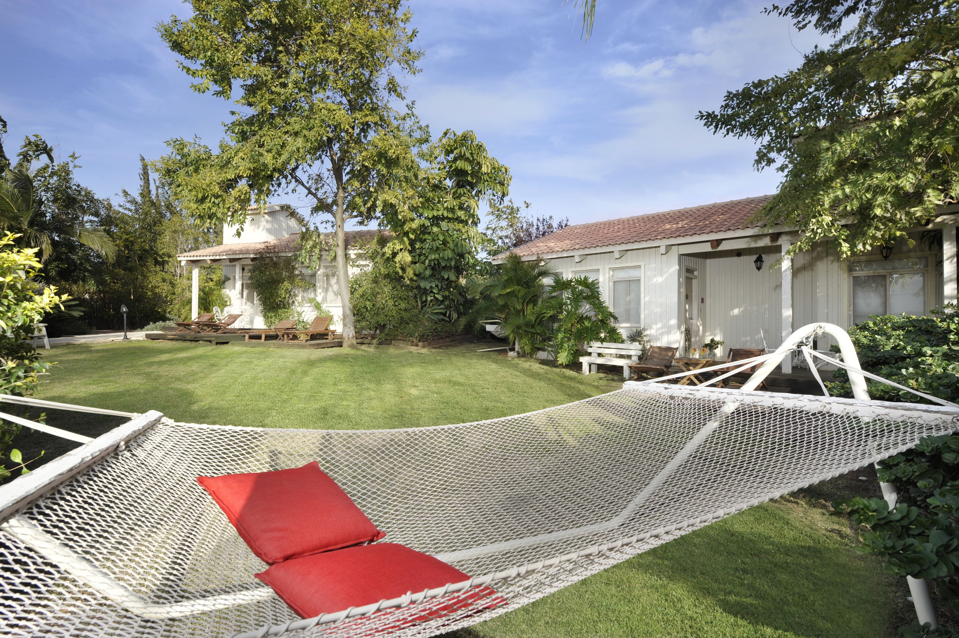 A hammock with red cushions in a grassy backyard with a white house and trees.