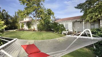 A hammock with red cushions in a grassy backyard with a white house and trees.