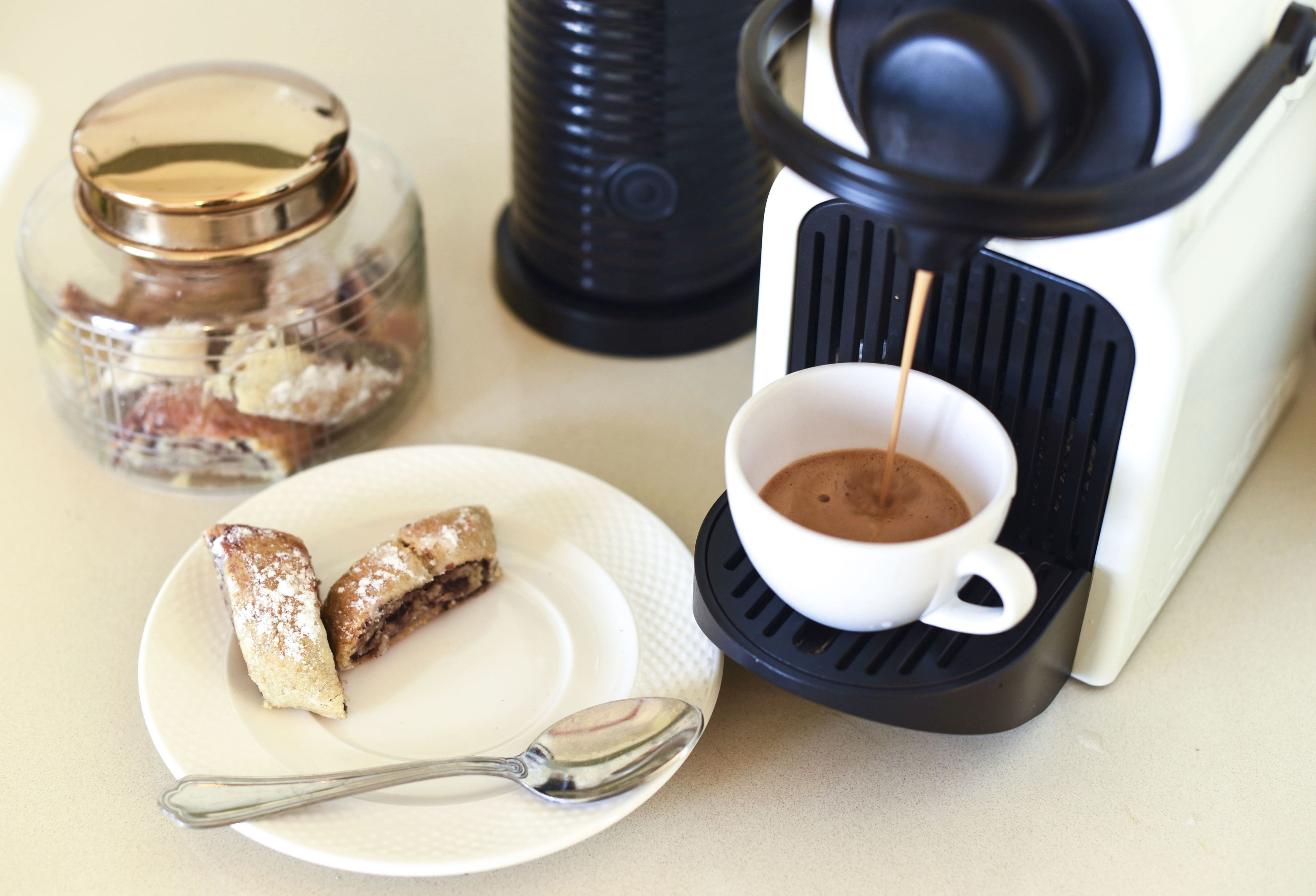 A coffee maker with a cup of coffee, a plate with pastries, and a jar of sugar.