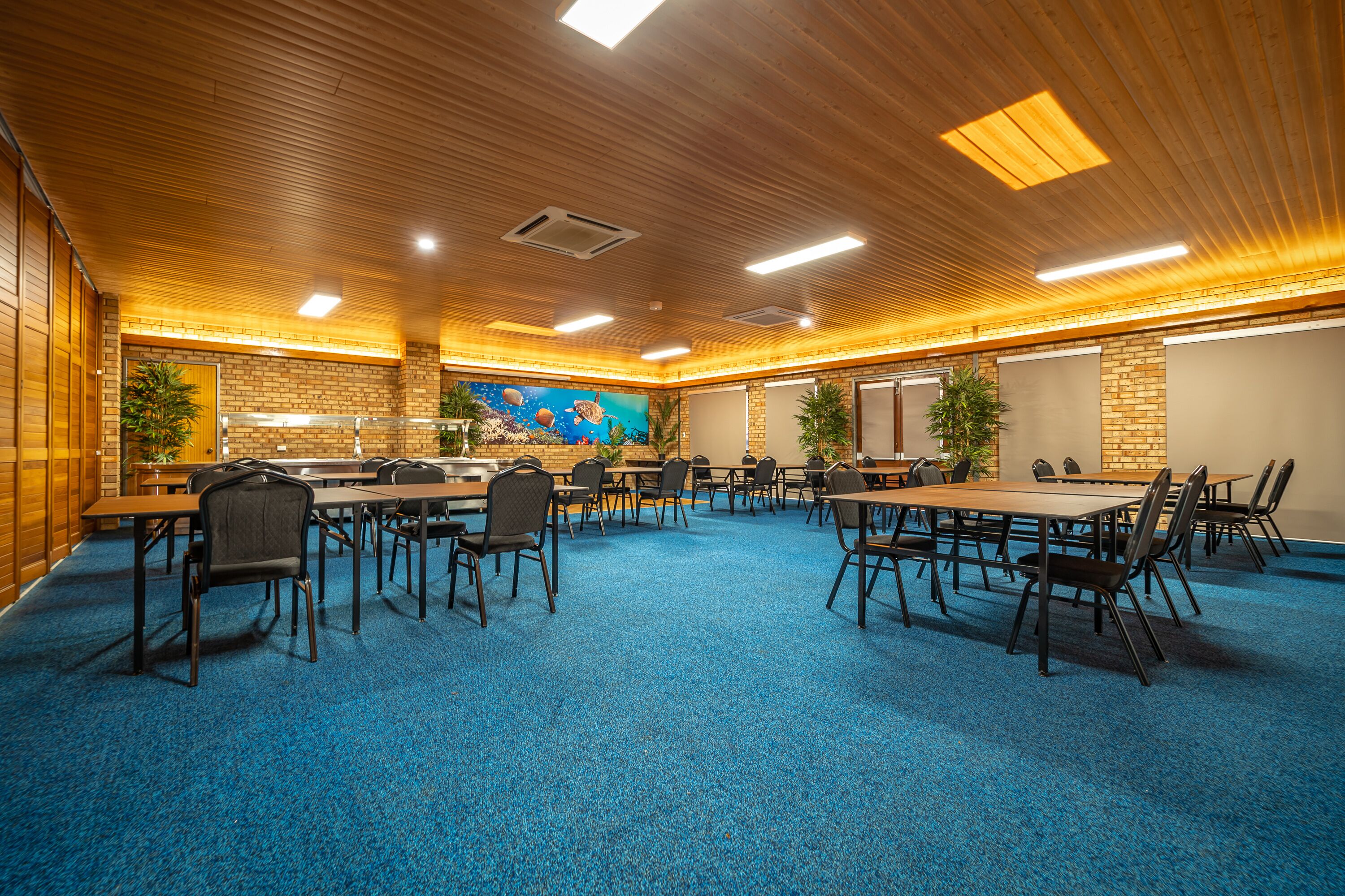 A conference room with a blue carpet, wooden ceiling, and brick walls. Tables and chairs are arranged for meetings.