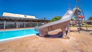 Une piscine avec un toboggan, entourée d’une zone délimitée par une clôture et avec un bâtiment visible au fond.