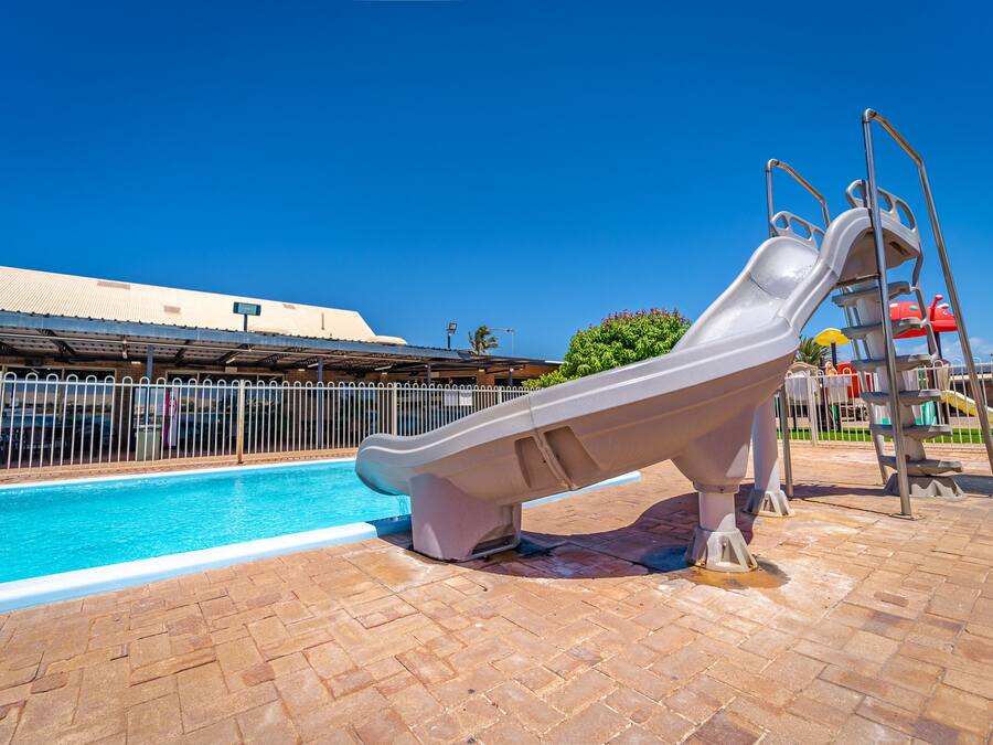 A swimming pool with a slide, surrounded by a fenced area and a building in the background.