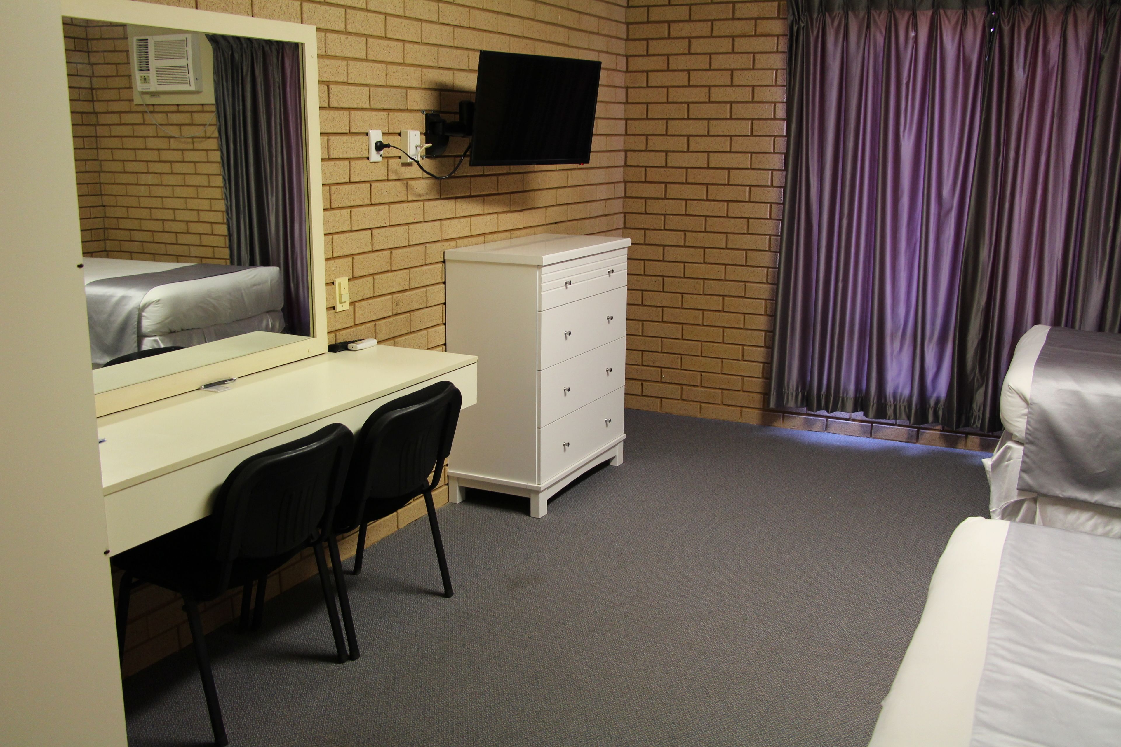 A hotel room with a bed, a desk with a chair, a dresser, a television mounted on the wall, and a window with curtains.