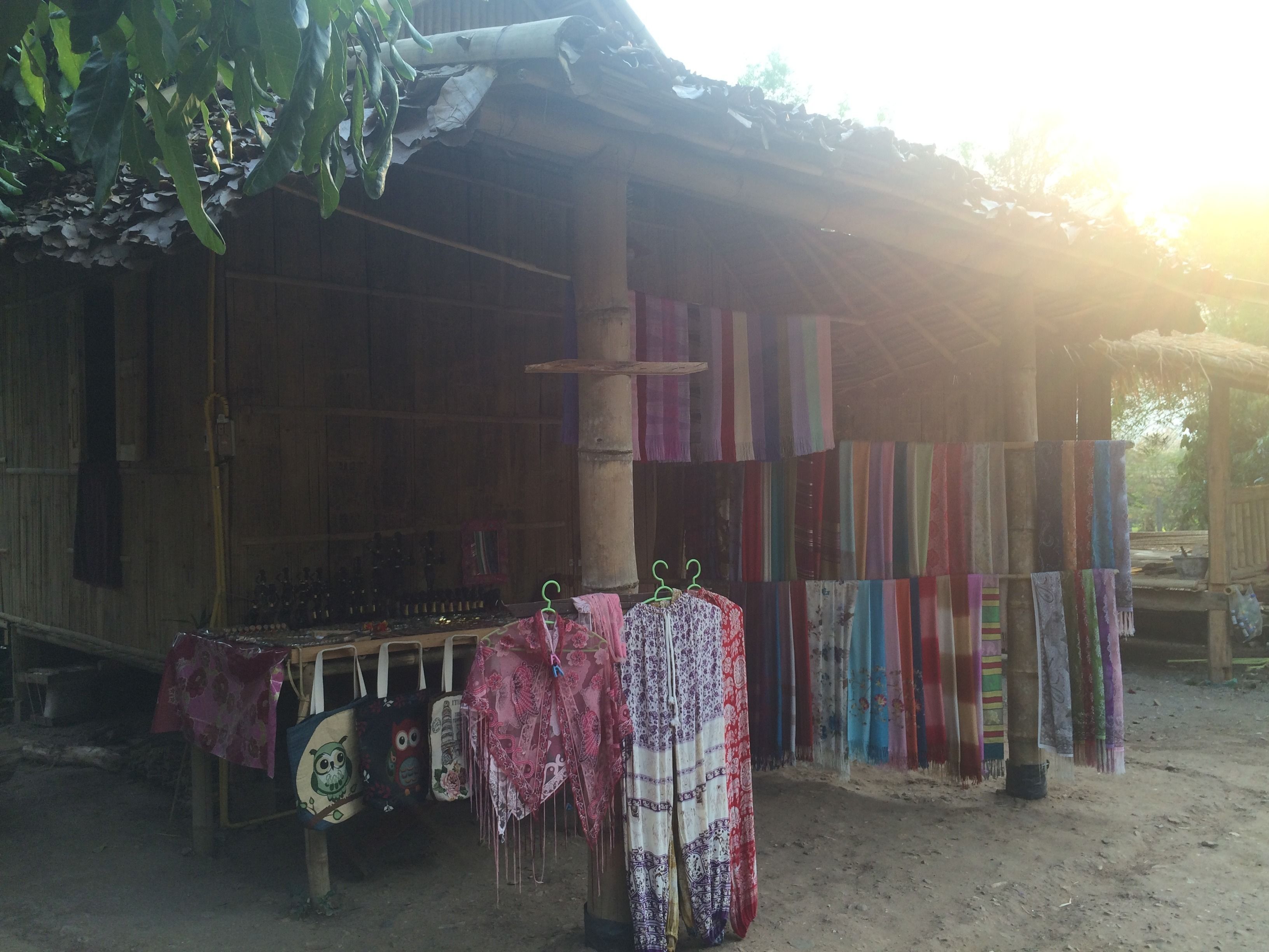 A rustic wooden structure with a thatched roof, displaying colorful fabrics and a variety of items on shelves.