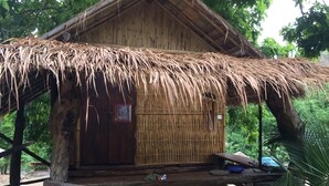 A rustic wooden hut with a thatched roof, a wooden door with a red sign, and a small porch area with a blue basin and a wooden bench. - Suan Pra - Inn (Pai)