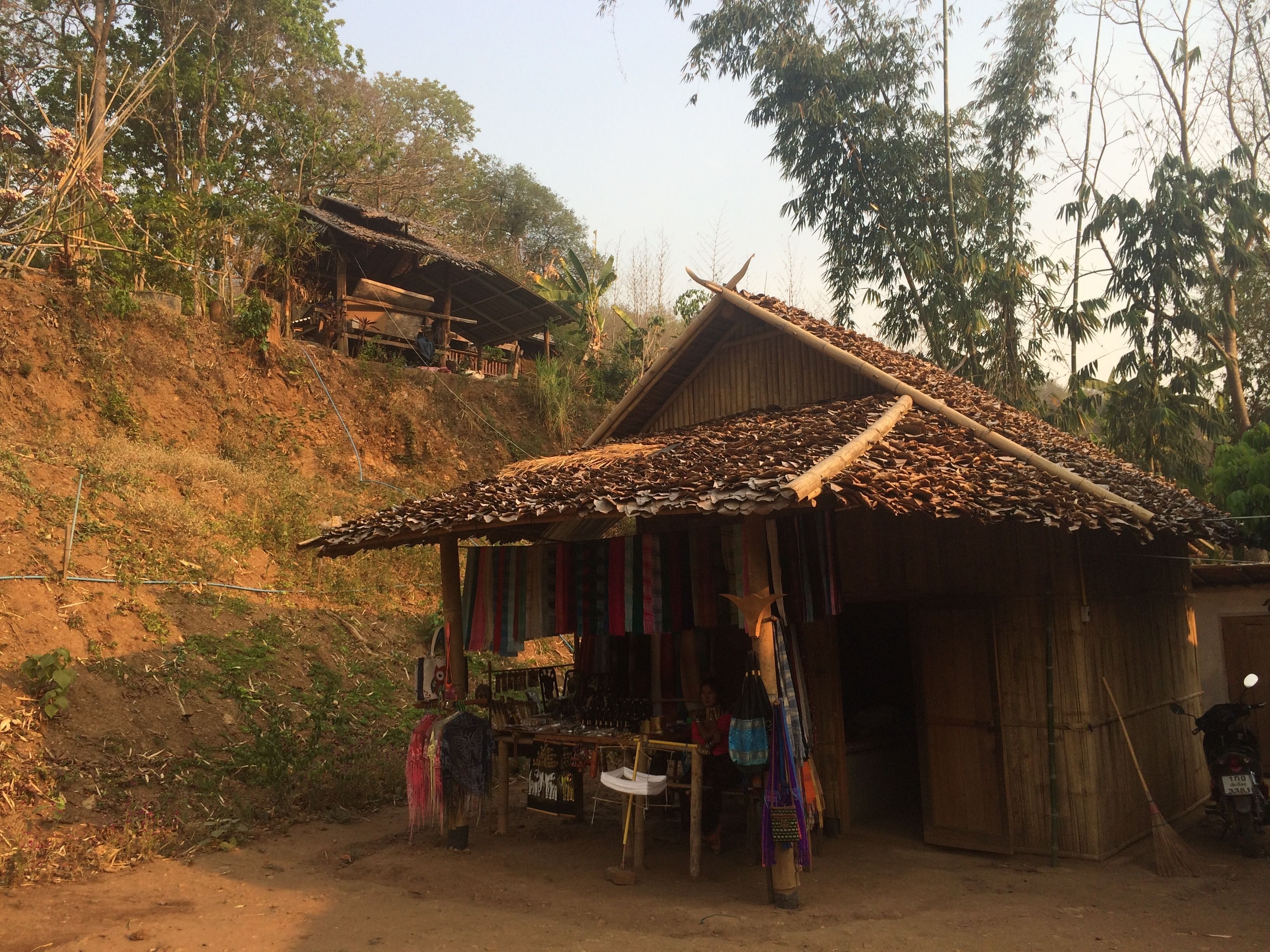 A rustic wooden structure with a thatched roof, a small shop with colorful items outside, and a dirt path leading to it.