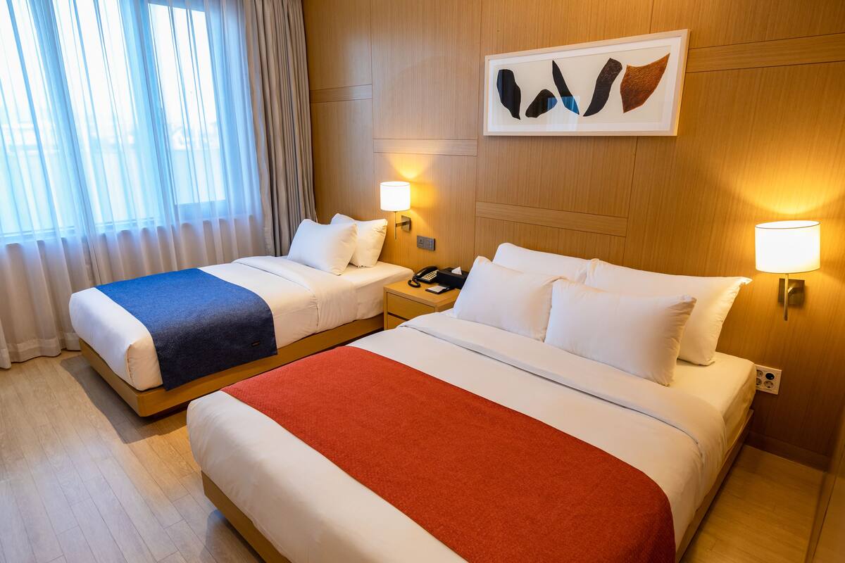 A hotel room with two beds, a large window with curtains, a wall-mounted artwork, and bedside lamps.
