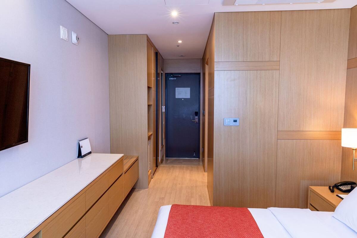 A hotel room with a bed, bedside table, TV, wardrobe, and a hallway leading to a door.