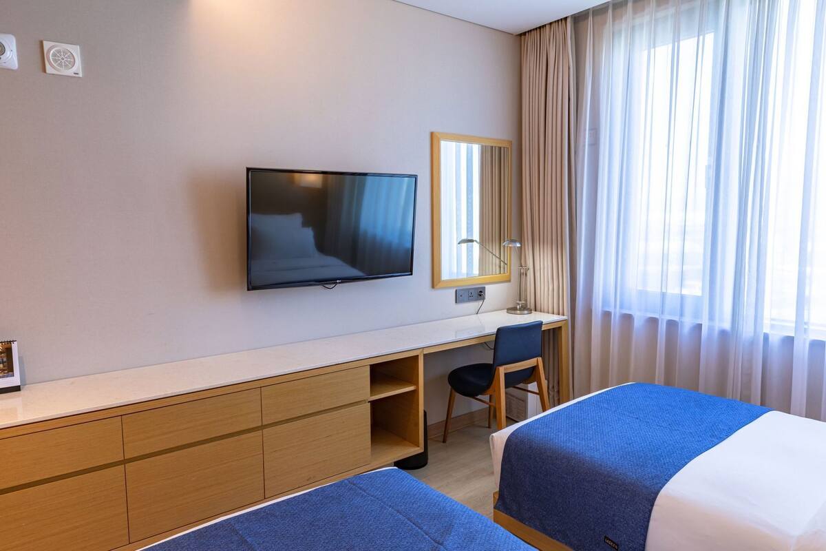 A hotel room with a bed, a desk with a chair, a television, a mirror, and a window with curtains.