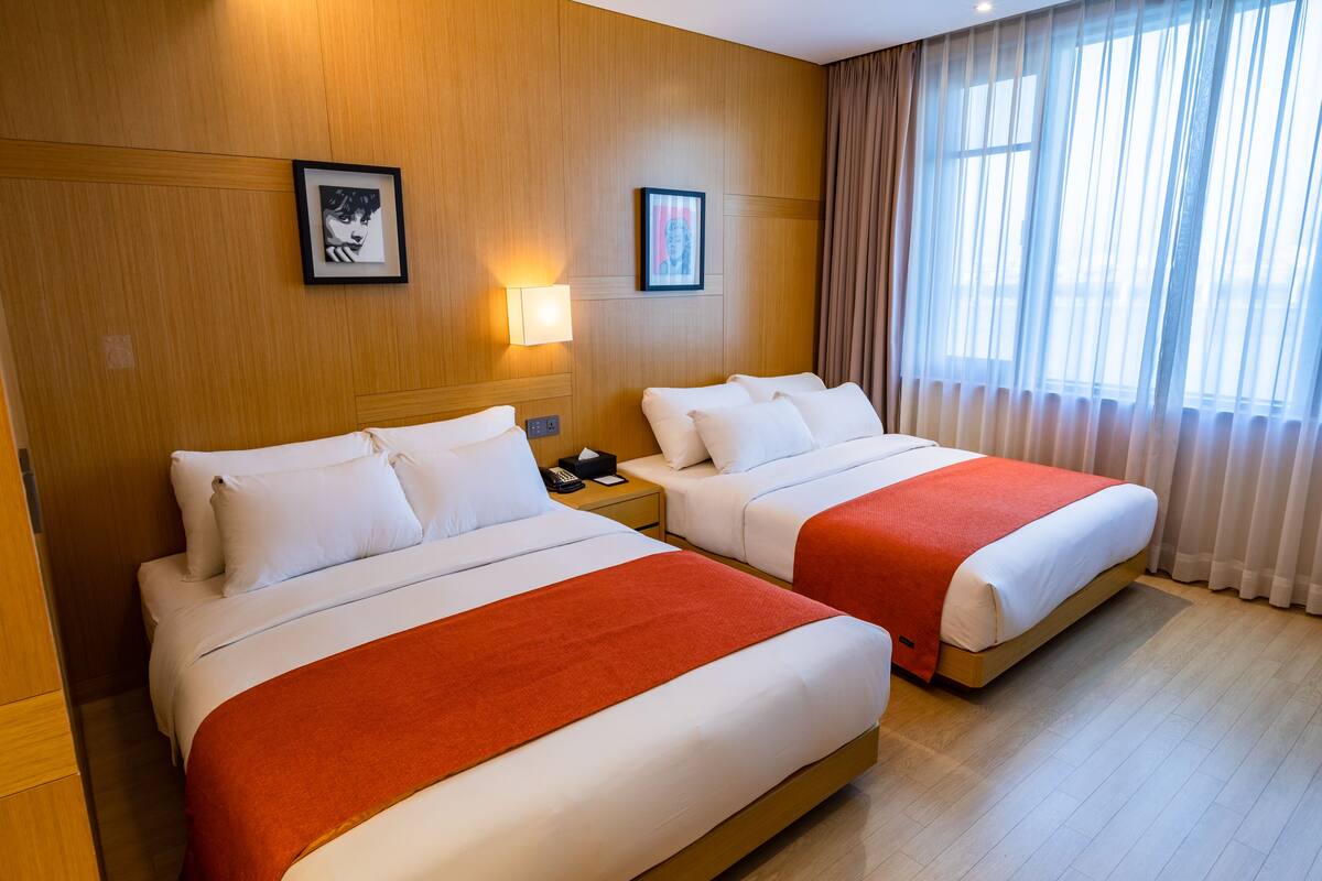 A hotel room with two beds, each with white bedding and a red blanket. There are framed pictures on the wall, a bedside lamp, and a window with curtains.