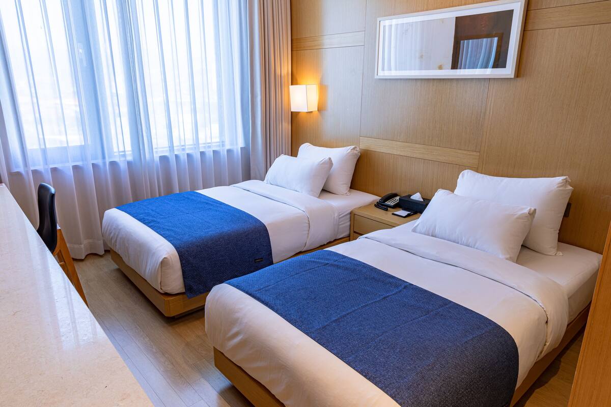 A hotel room with two beds, a desk, a chair, a nightstand with a phone, and a wall-mounted lamp.