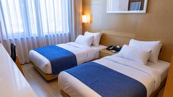 A hotel room with two beds, a desk, a chair, a nightstand with a phone, and a wall-mounted lamp.