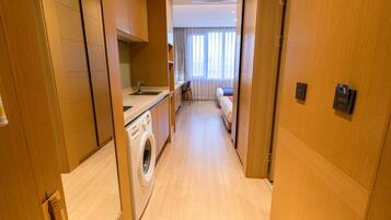 A hotel corridor with a washing machine, kitchenette, and a bed in the distance.