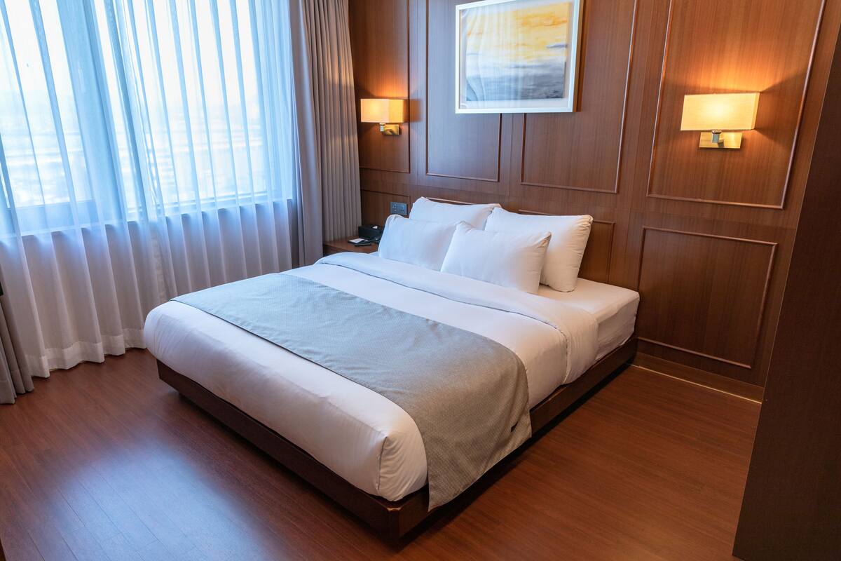 A hotel room with a large bed, wooden paneling, two bedside lamps, a painting on the wall, and a window with sheer curtains.