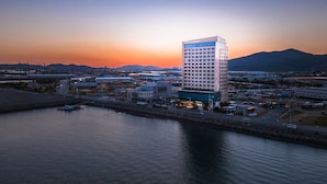 A modern hotel with a prominent tower, situated by a calm body of water, with industrial buildings and mountains in the background. - Hotel Lacky Gwangyang (Gwangyang)