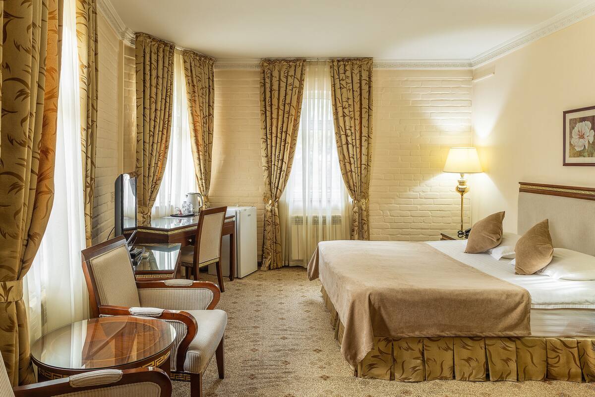 A hotel room with a bed, a desk with a chair, a lamp, and curtains.