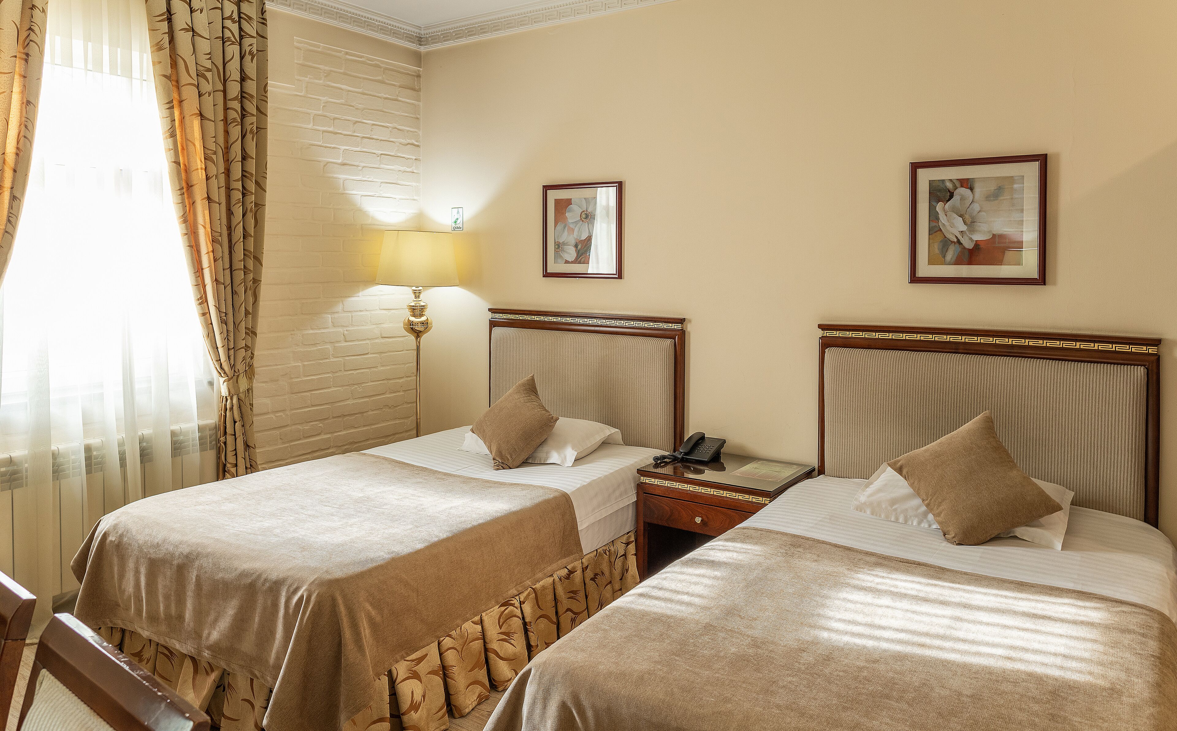 Quarto de hotel com duas camas de solteiro, mesa de cabeceira com uma lâmpada, telefone e dois quadros na parede.