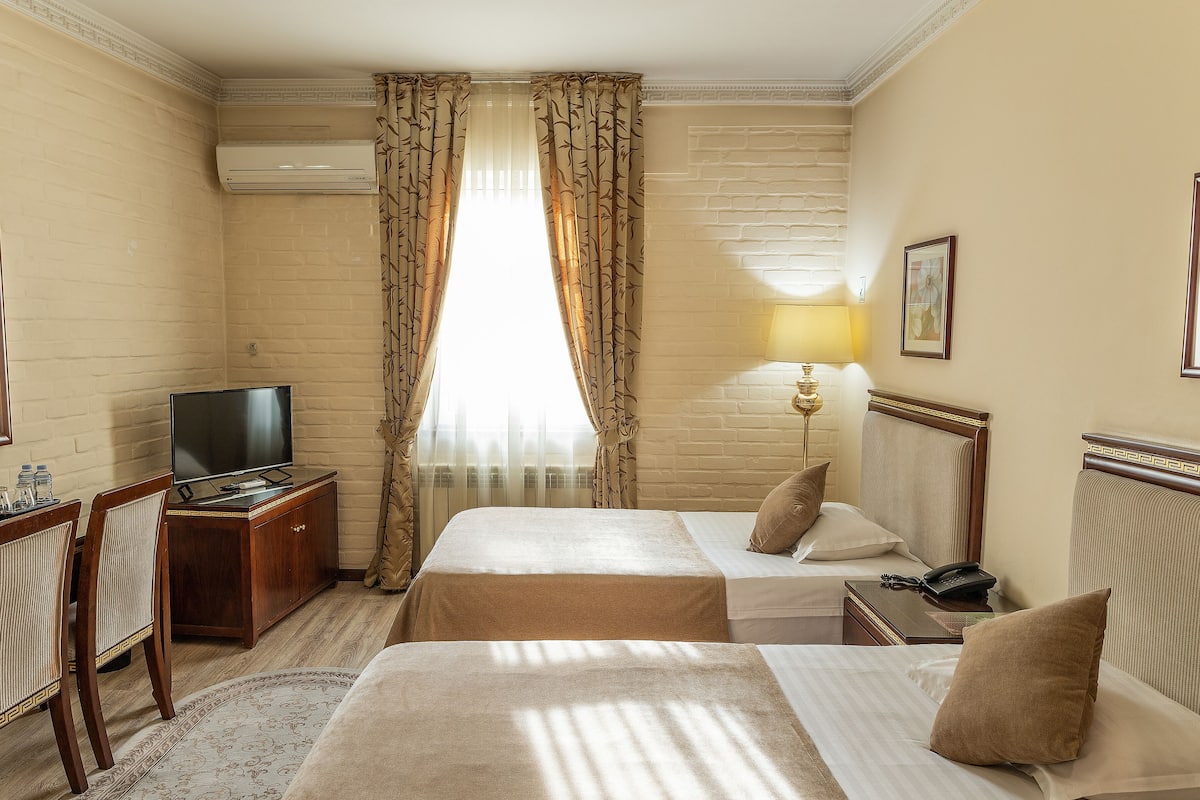 A hotel room with two beds, a desk with a TV, a lamp, and a window with curtains.
