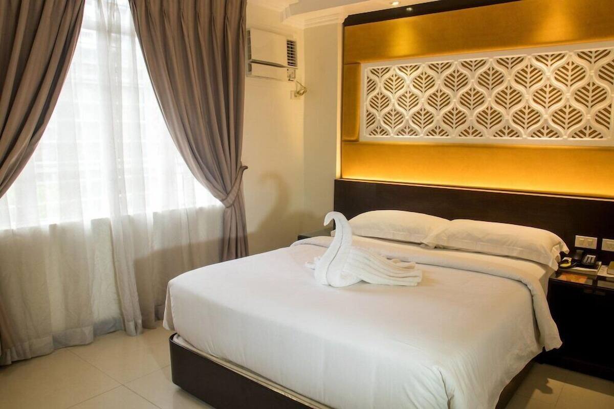 A hotel room with a bed, curtains, a wall-mounted air conditioner, and a decorative panel above the bed.