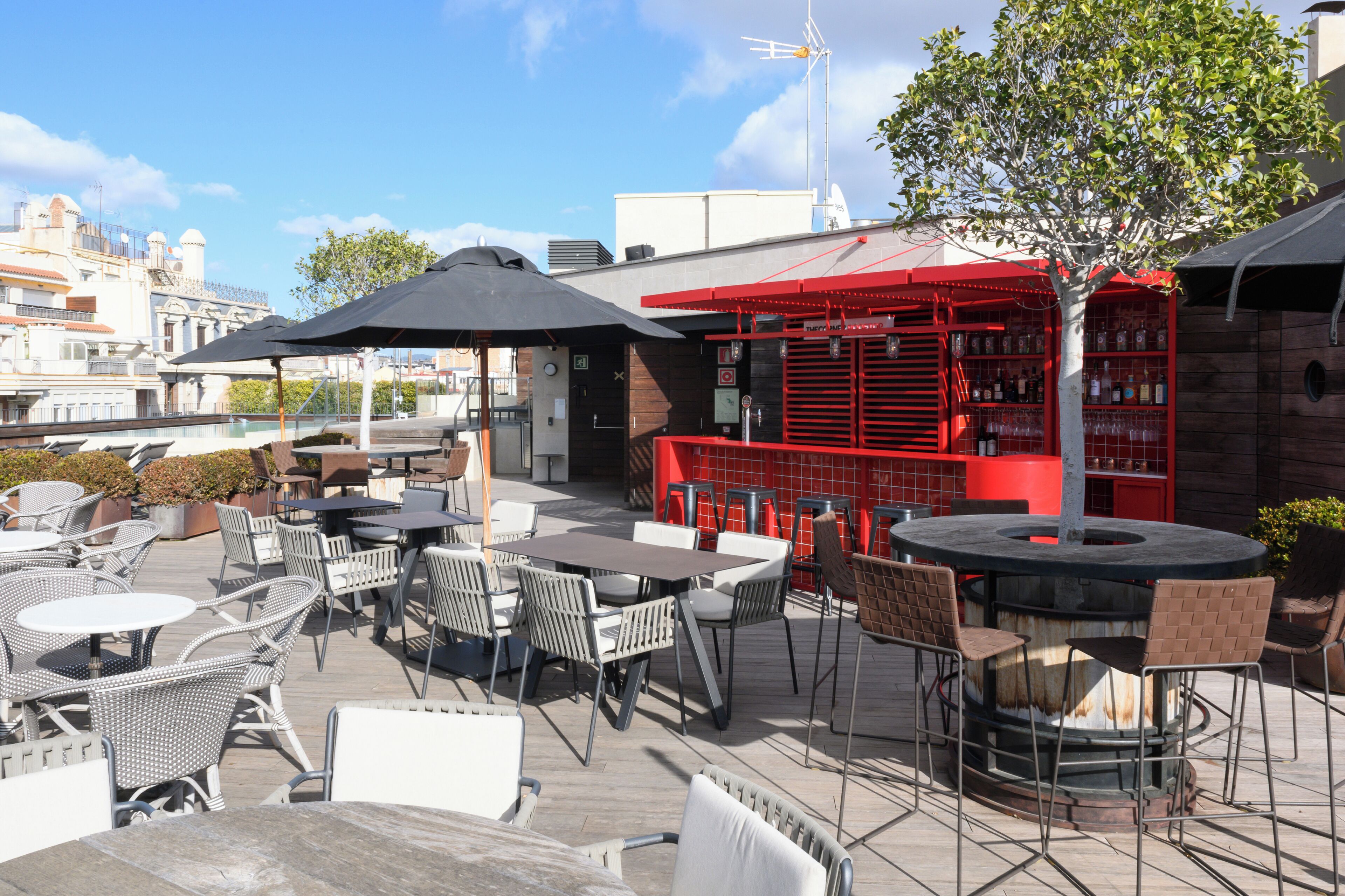 Een terras buiten met tafels en stoelen, een rode bar en gebouwen op de achtergrond.