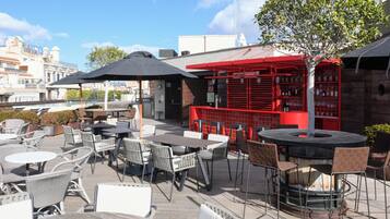 Een terras buiten met tafels en stoelen, een rode bar en gebouwen op de achtergrond.