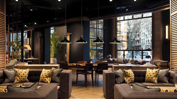 A modern interior with a central dining area, sofas with patterned cushions, and large windows providing a view of the city. - The Corner Hotel (Barcelona)
