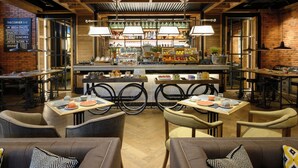 A restaurant with a brick wall, wooden ceiling, and tables with chairs. There are signs on the wall and a counter with food items. - The Corner Hotel (Barcelona)