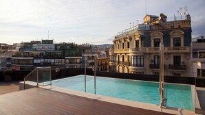 A rooftop swimming pool with a wooden deck, surrounded by buildings with balconies and a mix of architectural styles. - The Corner Hotel (Barcelona)