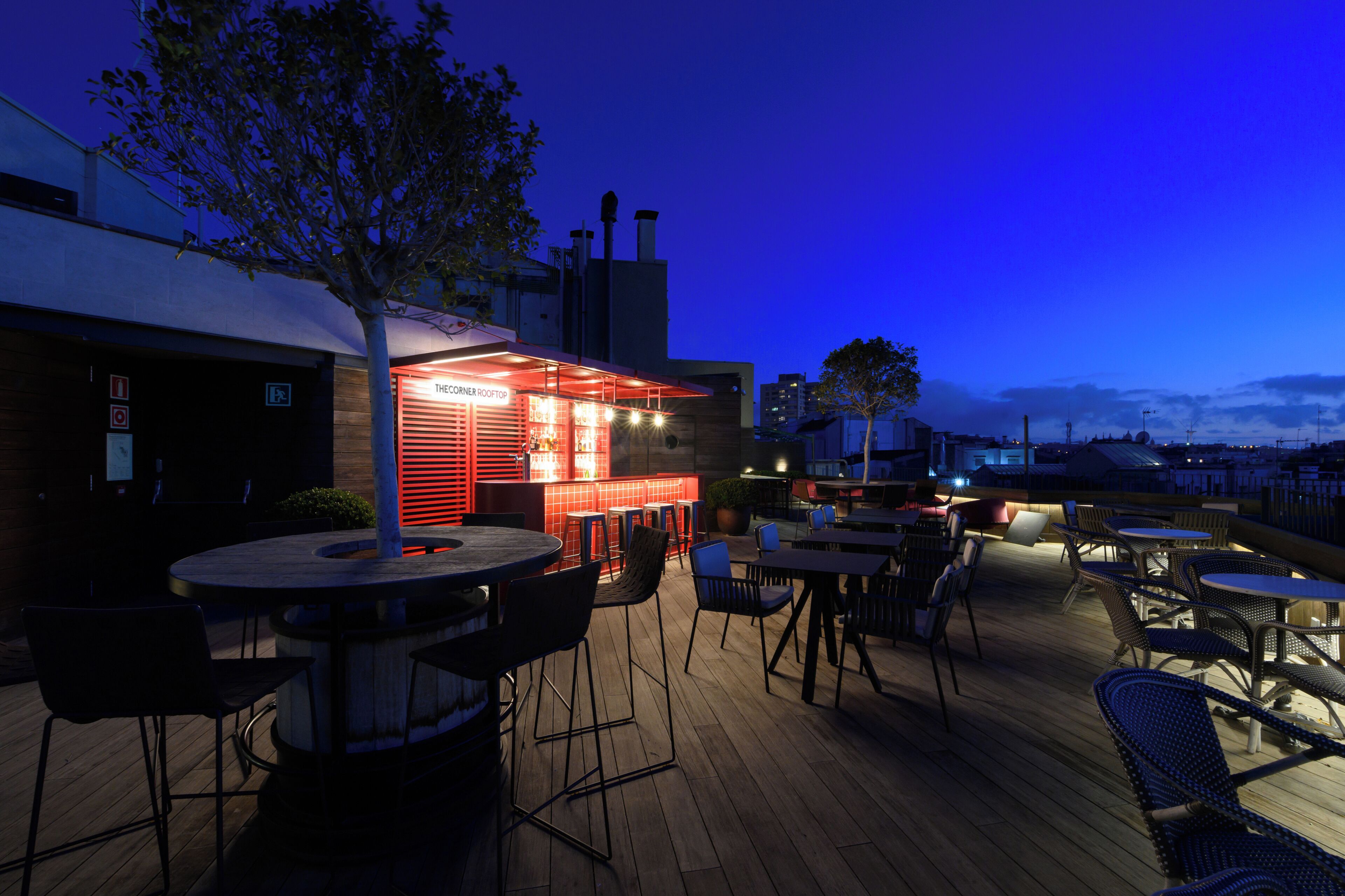 Un área de bar al aire libre con mesas y sillas, una barra iluminada en rojo y vistas del horizonte de la ciudad al anochecer.
