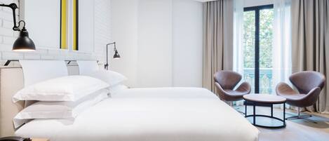 A hotel room with a large bed, two bedside lamps, a wooden nightstand, a telephone, a round table with two chairs, and a view of greenery through the window.