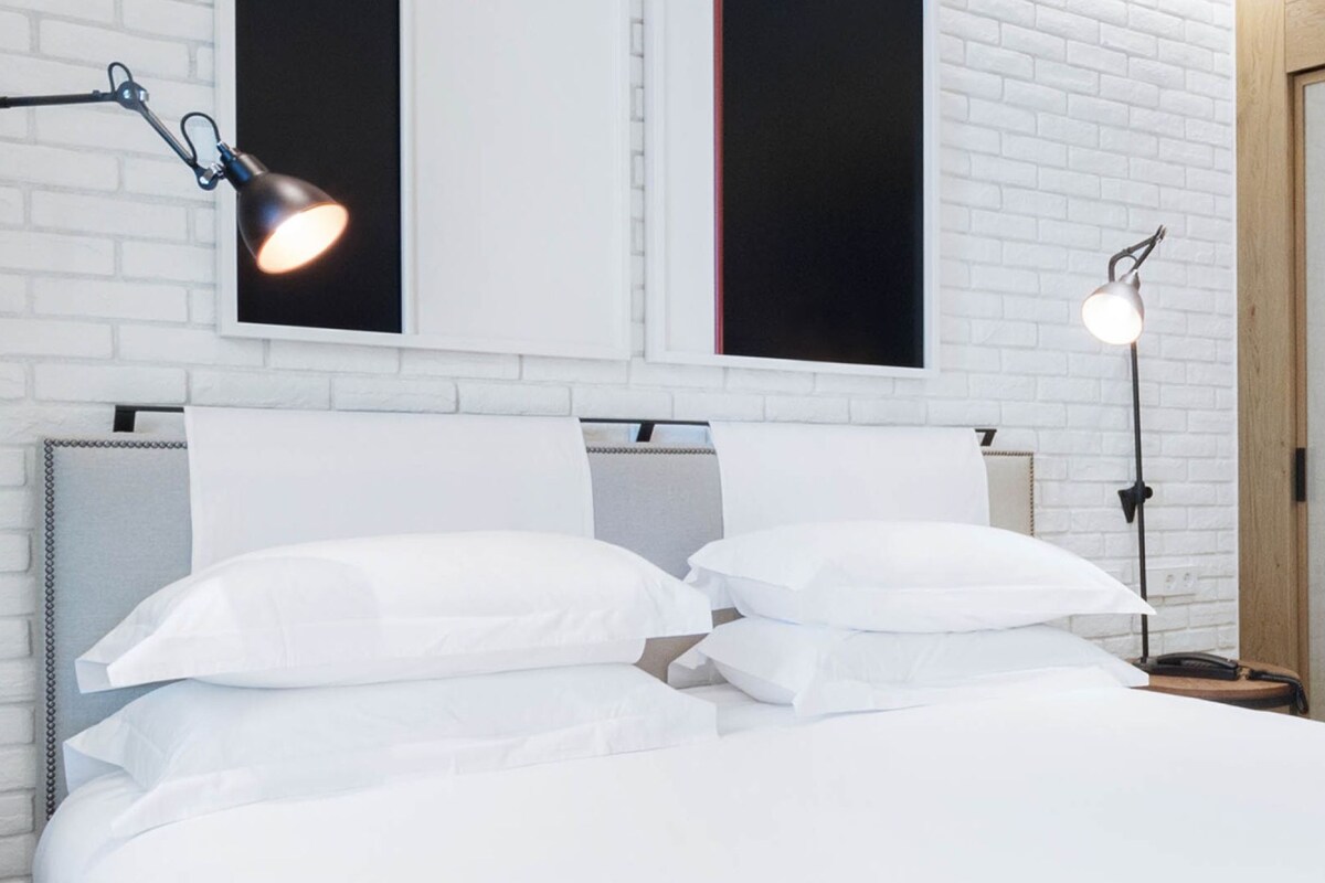 A modern bedroom with a white brick wall, a bed with white bedding, and two wall-mounted lamps.