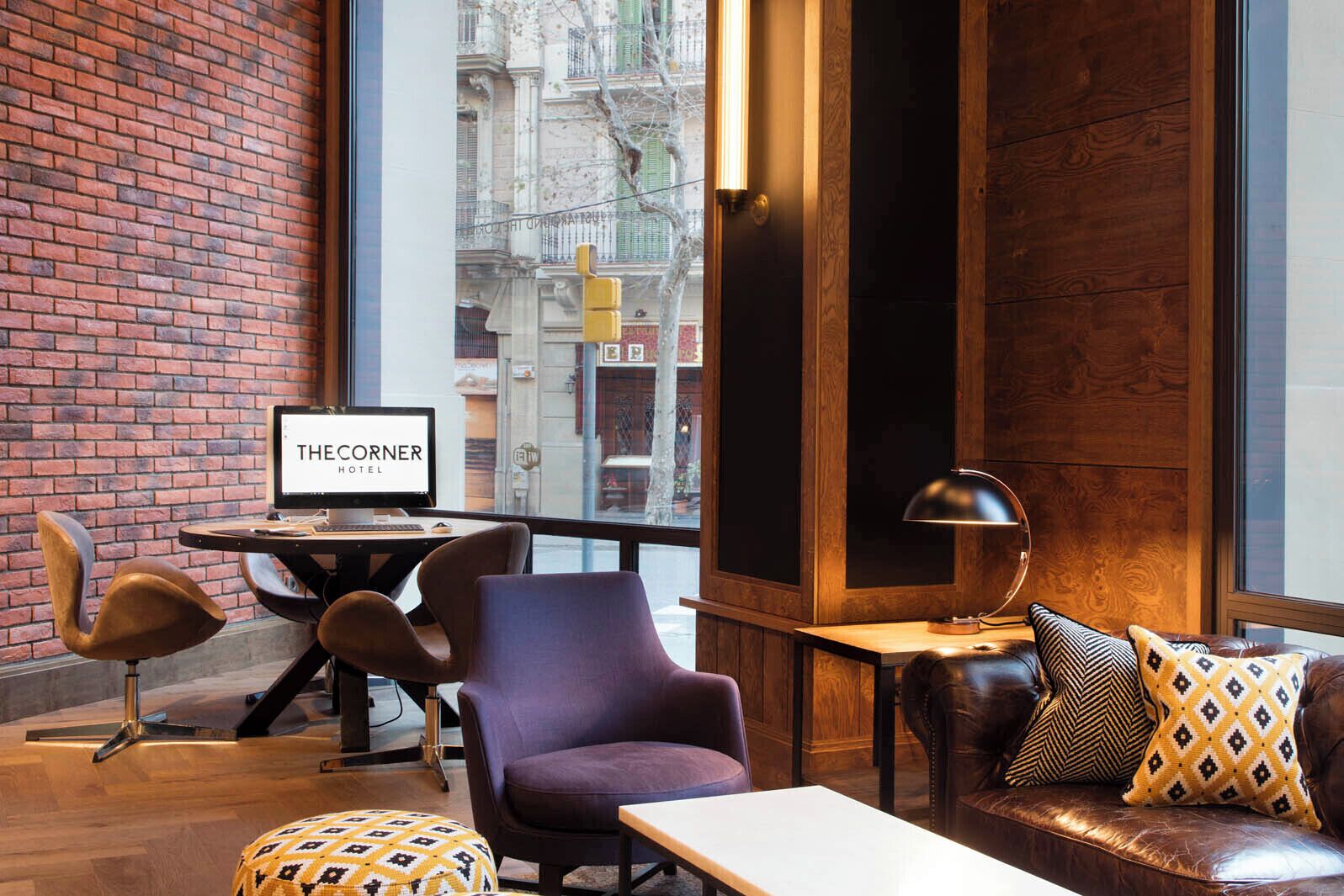 A hotel room with a brick wall, a wooden panel wall, a large window with a city view, a desk with a computer, a leather sofa, and patterned cushions.