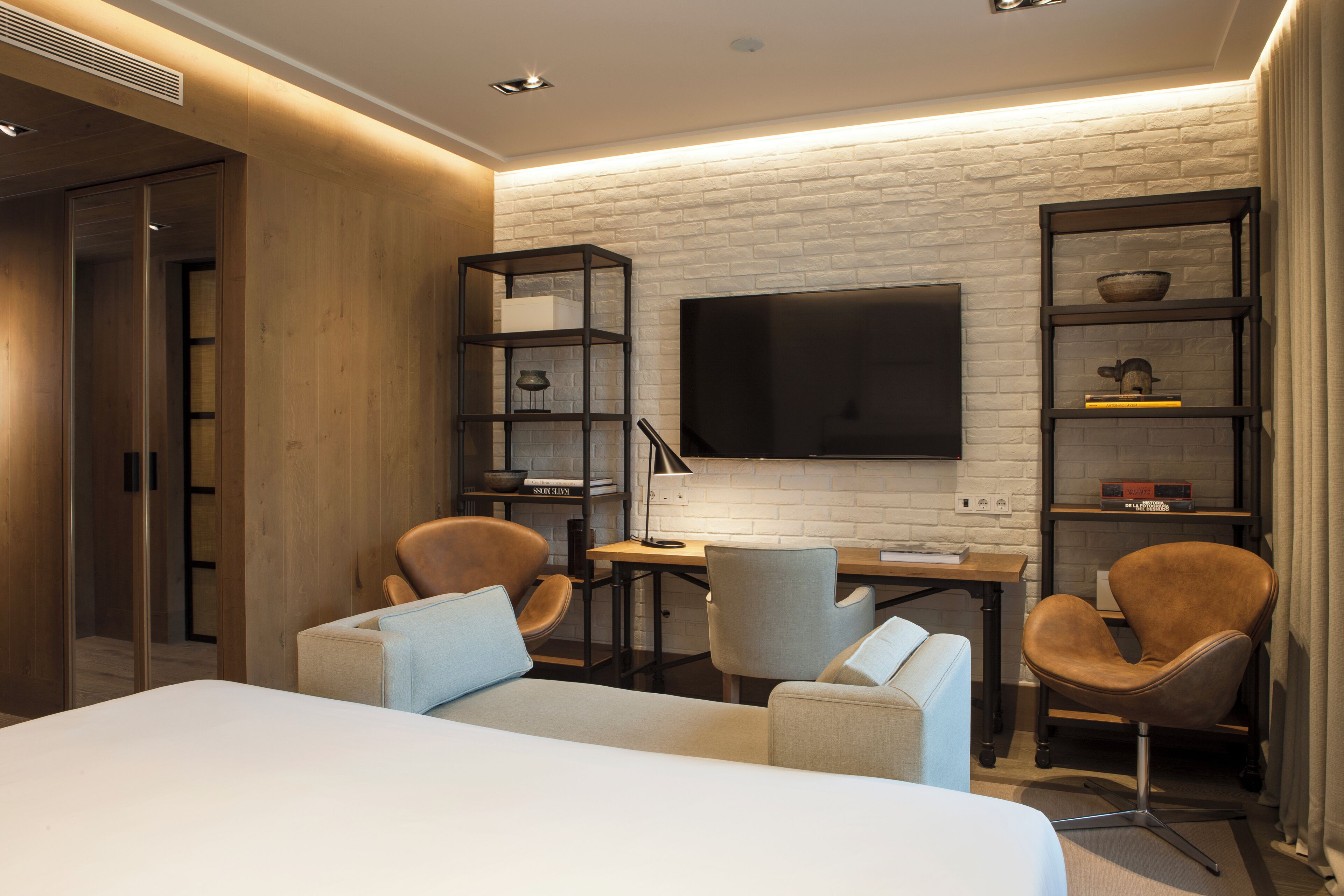 A modern hotel room with a large bed, two armchairs, a desk, and a TV mounted on a brick wall.