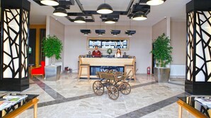 Un hall con mostrador de recepción, carrito de madera, plantas en macetas e iluminación colgante moderna.