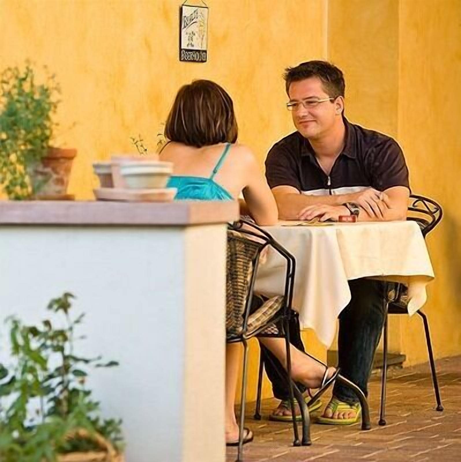 Two people seated at an outdoor table with a yellow wall in the background.