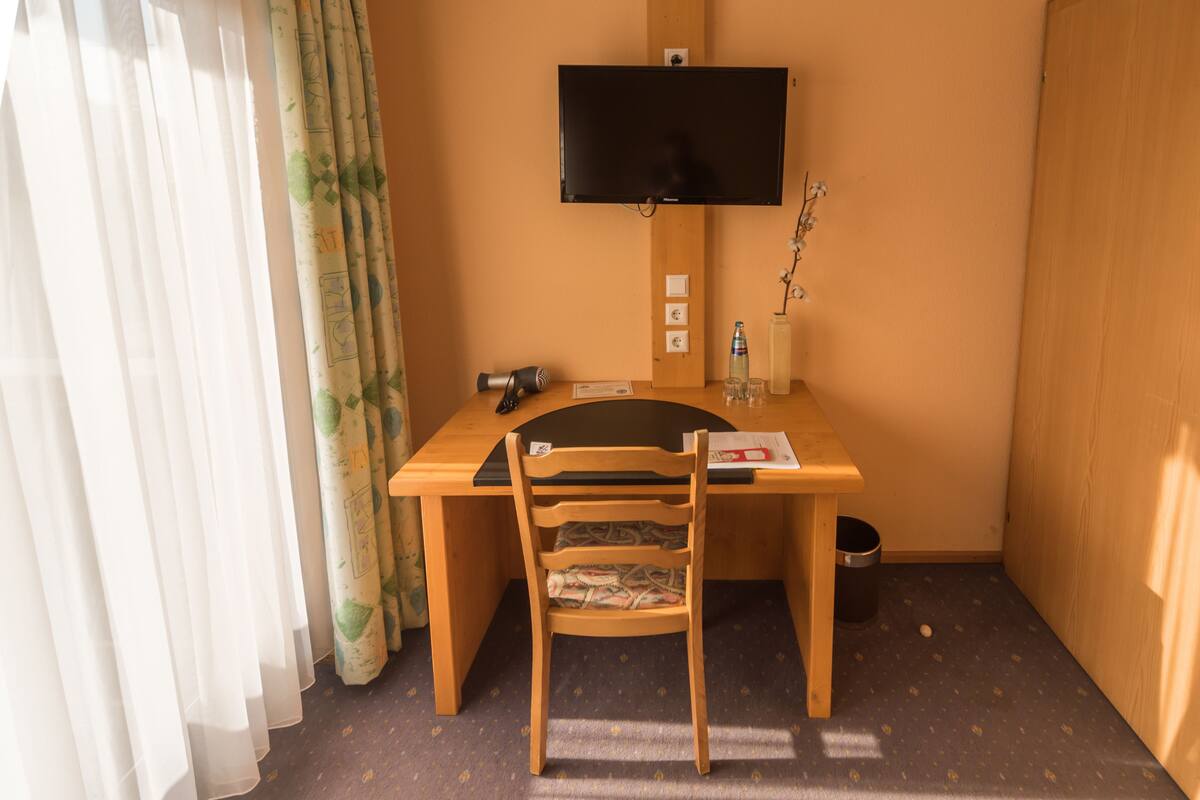 Una habitación de hotel pequeña con un escritorio de madera, una silla, un televisor montado en la pared, una ventana con cortinas ligeras y un cubo de basura pequeño.