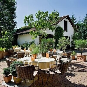 Une terrasse extérieure avec des tables rondes et des chaises en osier, entourée de plantes en pot et d’un bâtiment blanc en arrière-plan.