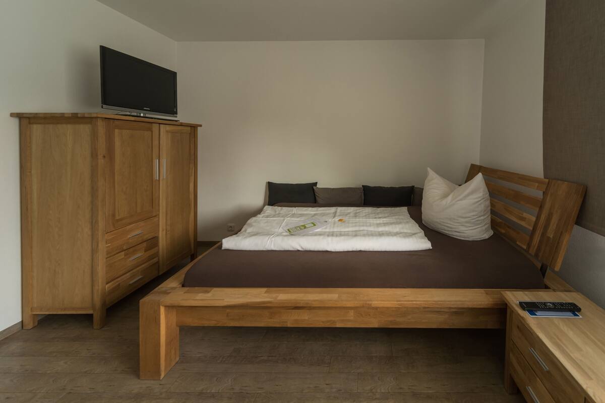 Un dormitorio con una cama de madera, un televisor sobre un mueble bajo, un armario de madera y una mesita de noche con un control remoto.