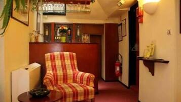 Hall d'albergo con una poltrona a scacchi, un tavolino, un'area bar con bottiglie e bicchieri, ed un estintore.