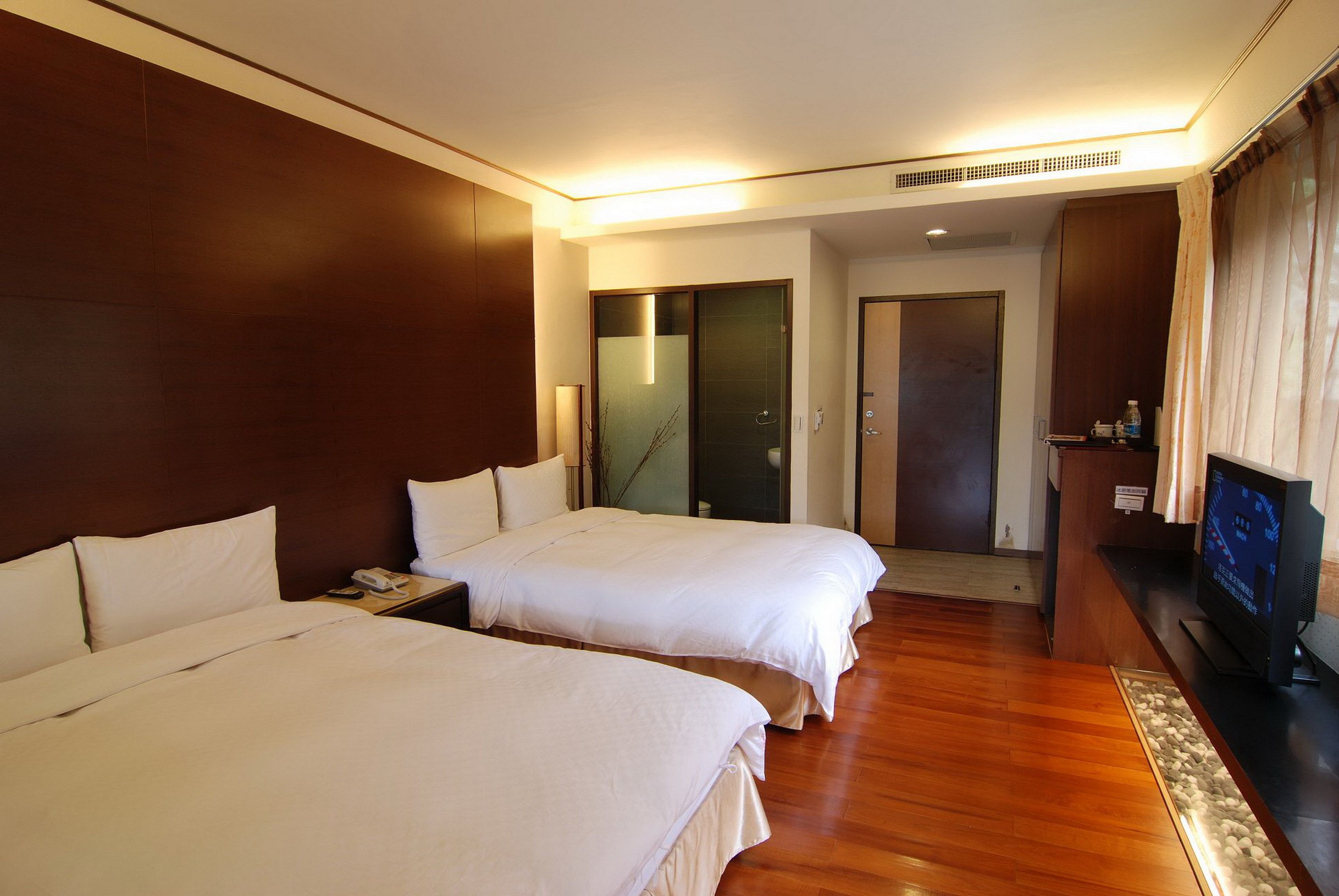 A hotel room with two beds, a television, a desk with a phone, and a wooden floor.