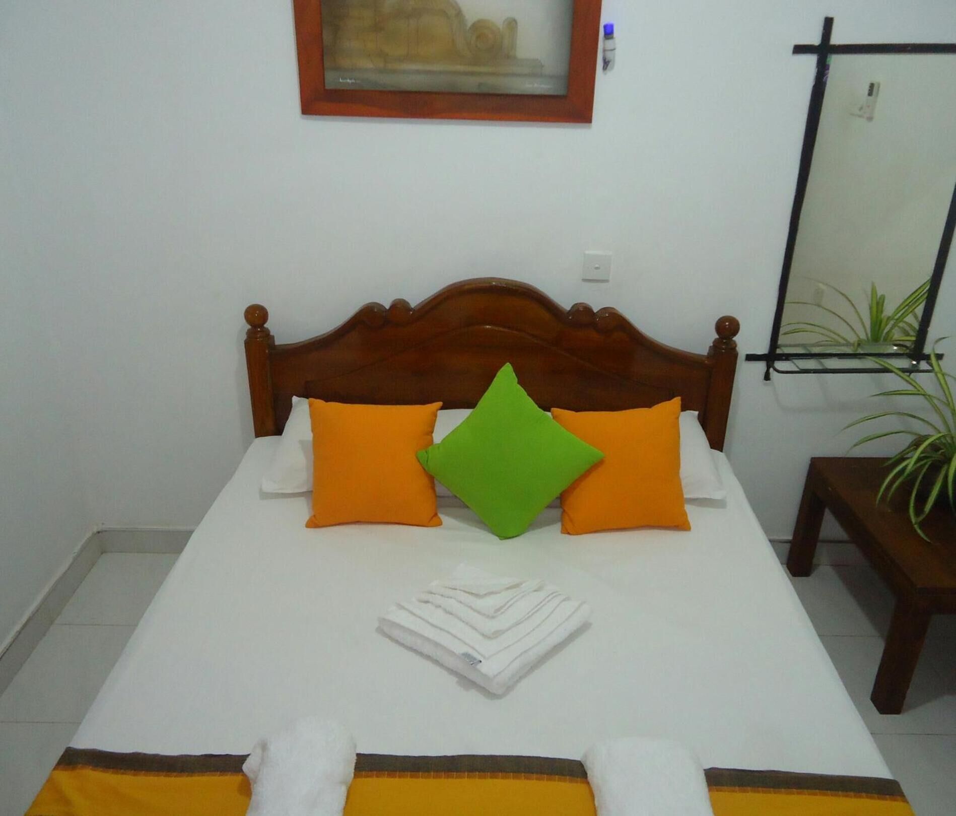 A neatly made bed with a wooden headboard, two orange pillows, one green pillow, and a folded white towel.