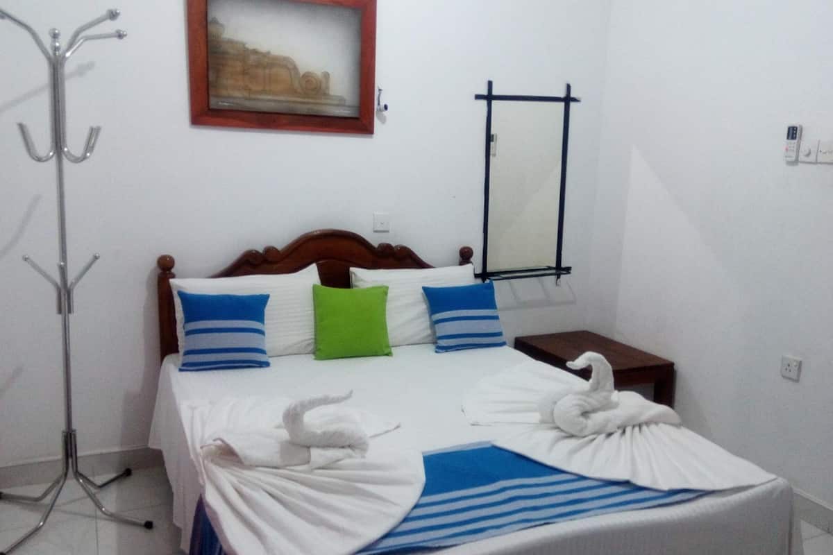 A neatly made bed with a wooden headboard, a framed picture above it, a mirror on the wall, and a coat rack to the left.