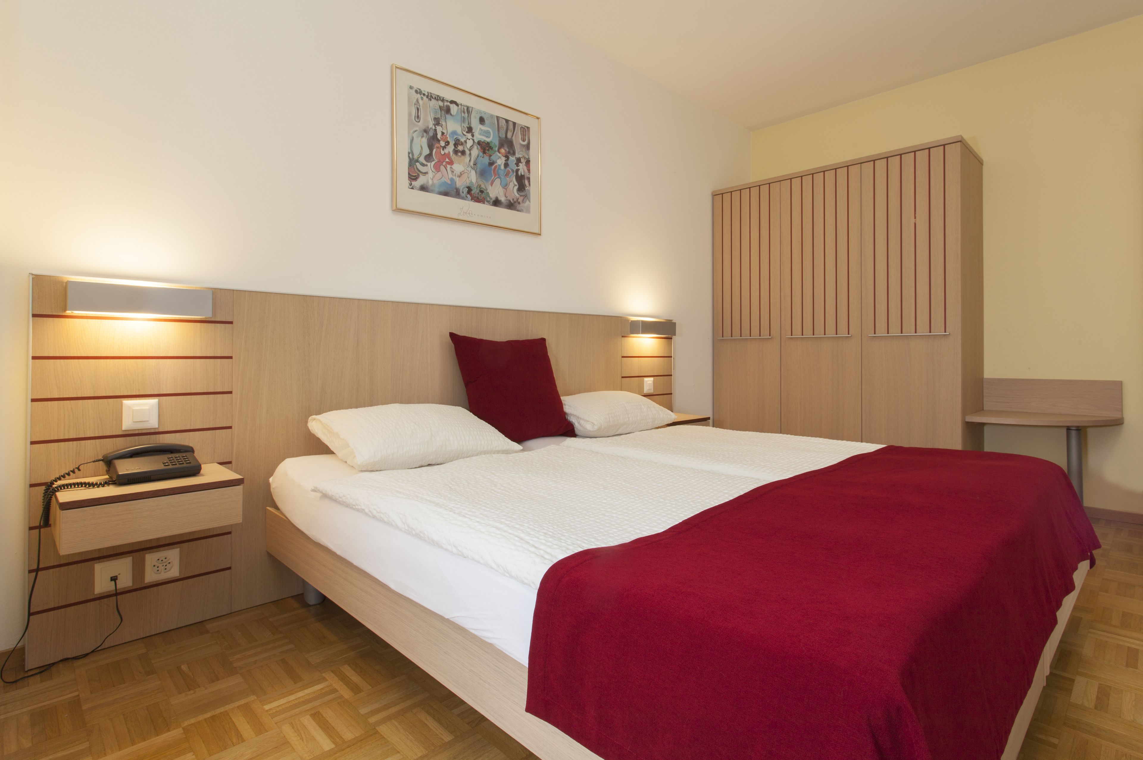 A neatly made bed with a red blanket, a bedside lamp, a telephone, and a wall-mounted artwork.