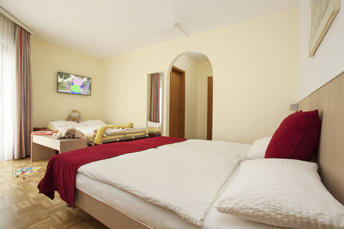 Habitación de hotel con una cama grande, un televisor en la pared, un escritorio con un osito de peluche y una almohada roja.