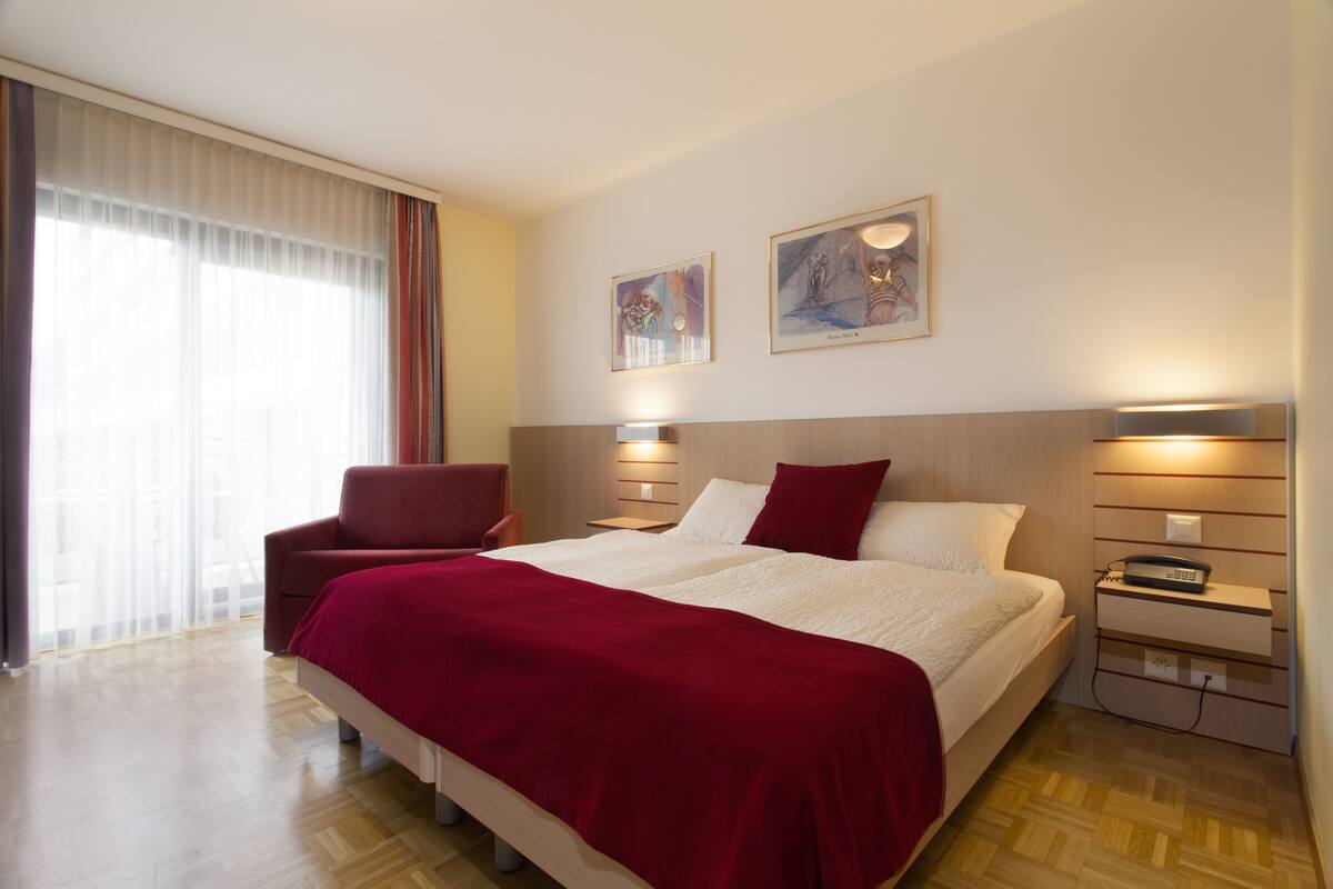 Habitación de hotel con una cama grande, una silla roja, una mesita de noche con una lámpara y dos cuadros en la pared.